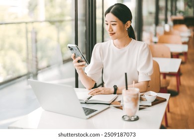 People relax at cafe concept. Happy face of adult asian woman using smart phone. On table with laptop computer and breakfast meal. Sitting near window indoors on day. - Powered by Shutterstock