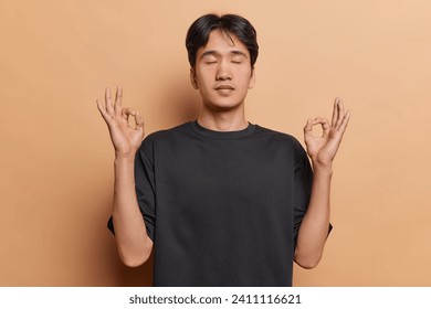 People and relaxation. Studio shot of young calm Asian man with closed eyes wearing black t shirt meditating after hard working day to relax and calm down standing isolated on beige backgorund - Powered by Shutterstock