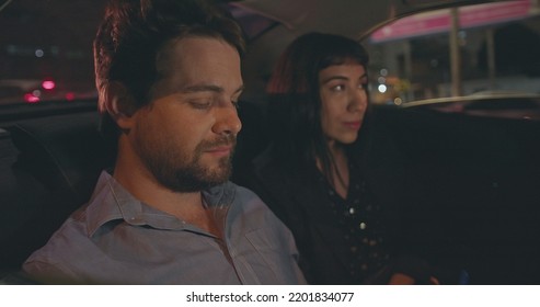 People Riding Taxi Cab In Car Backseat At Night. Young Man And Woman Passengers In The Evening After Work Talking On Phone