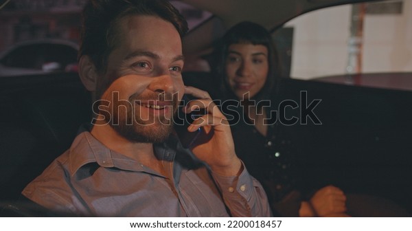 People riding taxi cab in car backseat at night.\
Young man and woman passengers in the evening after work talking on\
phone