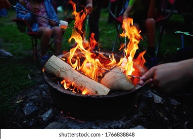 People Roasting Marshmallows On Skewers On Fire Pit At Campsite. Enjoying Summer Vacation Outdoor Camping Fun And Friend Togetherness Relaxing Time When Park And Campsite Reopen After Covid Lockdown.