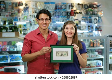 People Running Small Family Business, With Asian Shop Owner And Caucasian Wife In Computer Store, Showing Their First Dollar To The Camera And Smiling