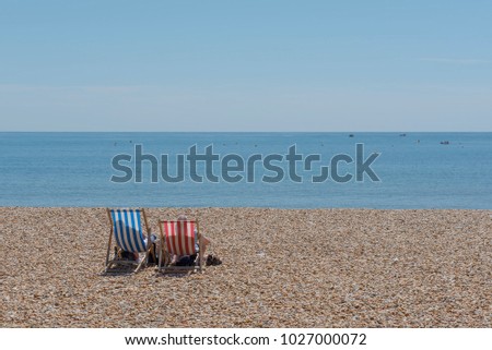 People sat in deck chairs  with shoes off on a pebble beach in Lyme Regis Dorset watching the boats on the sea under a clear blue sky.