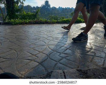 People are seen walking on a patterned concrete path, with a lush green park in the background