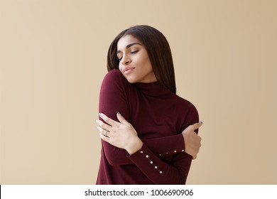People, self-love and self-esteem. Portrait of joyful attractive young dark skinned female closing eyes and hugging herself, having pleased relaxed facial expression. African girl embracing herself