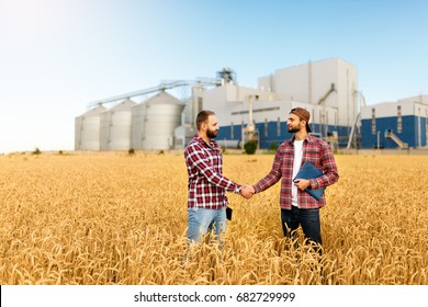 People shaking hands in a wheat field, farmer's agreement. Grain elevator terminal on background. Agriculture agronomist business contract concept