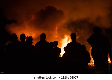 People Silhouetted Standing Around A Fire Set Off By Illegal Fireworks In California. A Bush Fire Outside Of A Building. 