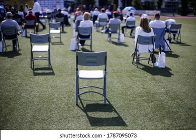 People Sit On Chairs Apart One From Another To Maintain The Social Distance During The Covid-19 Outbreak At An Outdoor Event On The Turf Of A Stadium.