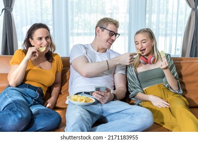 People Are Sitting On Coach And Having Snack For Dinner. Happy Family Eating Potato Ship And Watching Smartphone Together On Sofa At Home