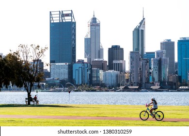 People Sitting On Park Bench And Cycling Girl At Sir James Mitchell Park In South Perth, With The Swan River And Perth City Skyline In Shot. PERTH, WESTERN AUSTRALIA. April 25, 2019.