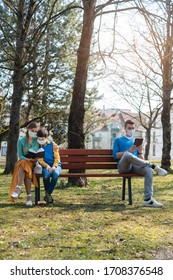 People Sitting On Park Bench In The Sun Practicing Social Distancing In Corona Crisis Wearing Masks