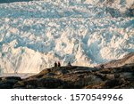 People sitting standing in front of huge glacier wall of ice. Eqip Sermia Glacier Eqi glacier in Greenland called the calving glacier during midnight sun. Hikers during travel and vacation.