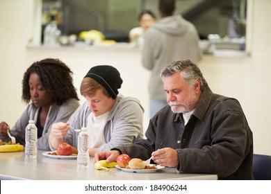 People Sitting At Table Eating Food In Homeless Shelter