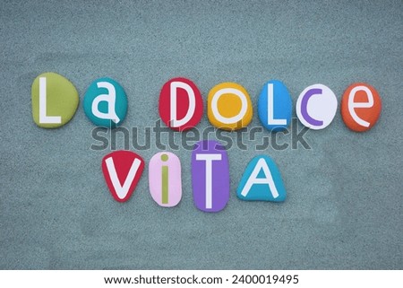 People sometimes use la dolce vita or the dolce vita to mean a life that is full of pleasure and luxury, creative text composed with multi colored stone letters over green sand