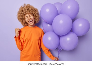 People special occasion and celebration concept. Optimistic woman wears orange jumper clenches fist celebrates special occasion holds bunch of inflated balloons isolated over purple background.