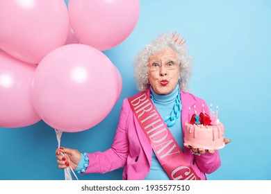 People Special Occasion Old Age Concept. Good Looking Senior Wrinkled Woman Keeps Lips Rounded Celebrates 102nd Birthday Poses With Birthday Cake And Bunch Of Balloons Isolated Over Blue Background