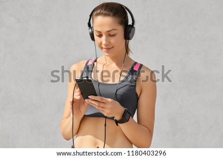 People, sport, music concept. Pleasant looking woman with healthy skin, wears casual top, smartwatch, concentrated inn screen of smart phone, listens favourite song in headphones, likes gymnastics