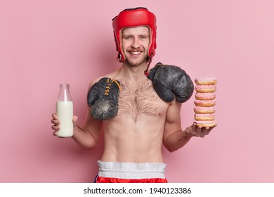 People Sport And Nutrition Concept. Positive Skinny Male Boxer Poses With Naked Torso Smiles Gladfully Has Temptation To Eat Donuts And Drink Milk Isolated Over Pink Background Holds Junk Food