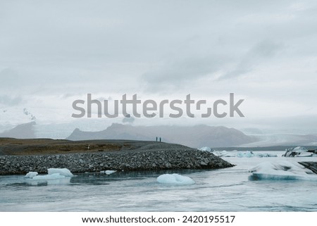 People stand on black sand hillside in rural Iceland overseeing glacial lake with floating icebergs, natural calm blue water, and snow capped mountains 