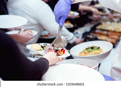 People Are Standing In Long Line In A Reustarant Buffet, Waiter Is Serving Food, Putting On Salad On A Plate With Tongs, Close Up Hands Concept 