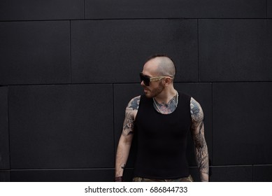 People, style and fashion concept. Attractive macho man with bald head and tattoos all over his arms and chest wearing black trendy sunglasses and tank top posing outdoors at blank copy space wall - Powered by Shutterstock