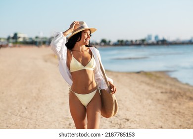 people, summer holidays and leisure concept - happy young woman in bikini swimsuit, white shirt and straw hat with bag walking along beach