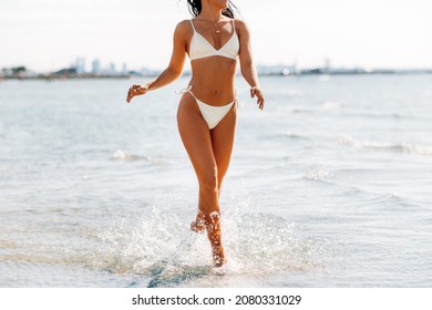 people, summer and swimwear concept - happy young woman in bikini swimsuit running in water beach