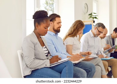 People take notes at business conference. Multiracial male female audience sitting in line in office room, listening to lecture and writing down information on clip boards, notepads, folders, binders