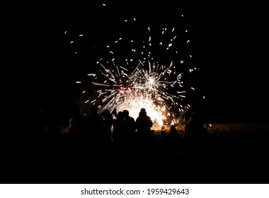 People take selfies and watch fireworks on beach with explosions of color against solid black night.