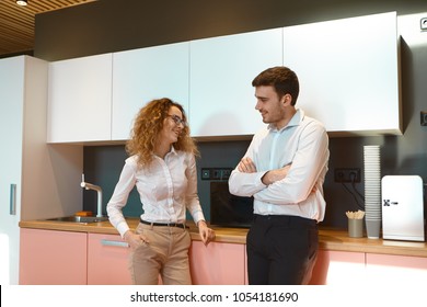 People, Teamwork, Cooperation, Career And Job Concept. Beautiful Young Woman Manager In Eyewear Standing In Office Kitchen And Flirting With Her Attractive Male Colleague, Both Smiling Playfully