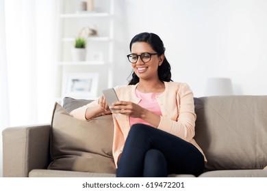 People, Technology, Communication And Leisure Concept - Happy Young Indian Woman Sitting On Sofa And Texting Message On Smartphone At Home