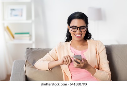 People, Technology, Communication And Leisure Concept - Happy Young Indian Woman Sitting On Sofa And Texting Message On Smartphone At Home