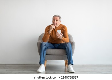 People And Technology Concept. Portrait Of Pensive Mature Man Using Phone And Thinking Looking Up, Sitting On Chair Isolated On White Studio Background Wall, Free Copy Space Mockup, Full Body Length