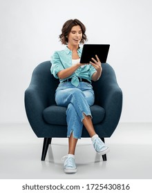 people and technology concept - portrait of smiling young woman in turquoise shirt and jeans with tablet pc computer sitting in modern armchair over grey background