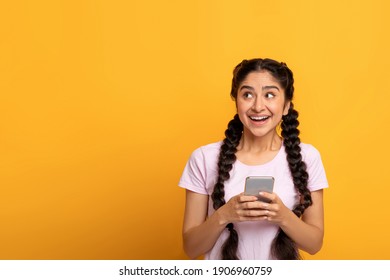 People And Technology Concept. Portrait Of Smiling Excited Attractive Indian Woman Using Smart Phone Standing Isolated On Yellow Orange Studio Background And Looking Aside At Copy Space, Banner