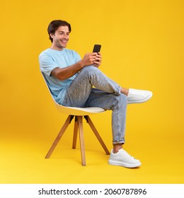 Gente Y Concepto De Tecnología. Retrato de un joven sonriente usando un smartphone sentado en una silla aislado en un estudio de color naranja. Persona casual emocionada chateando en línea, navegando por los medios sociales