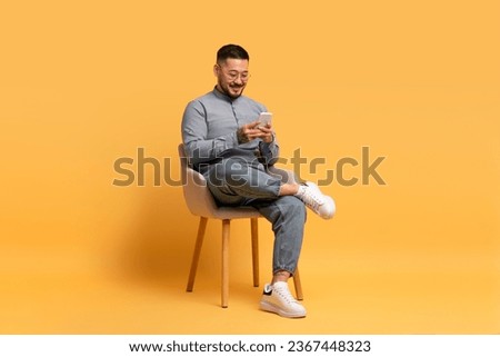 People And Technology Concept. Portrait of smiling asian man using smartphone while sitting on chair over yellow studio background. Excited casual guy chatting online, browsing social media