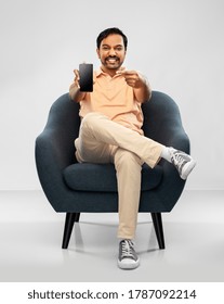 people, technology and furniture concept - happy smiling young indian man showing smartphone sitting in chair over grey background - Shutterstock ID 1787092214