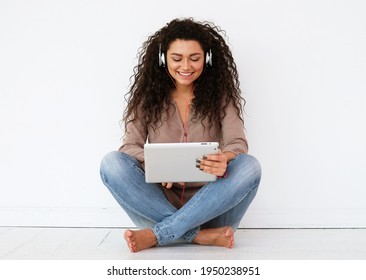 People, Technology And Leisure Concept - Happy African American Young Woman Sitting On Floor With Tablet Pc Computer And Headphones Listening To Music At Home