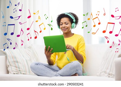 gente, tecnología y concepto de ocio - feliz joven afroamericana sentada en un sofá con tablet pc computadora y audífonos escuchando música en casa sobre notas de fondo