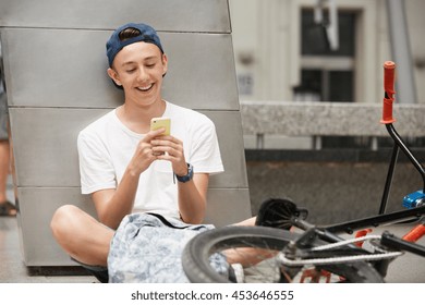 People, technology, leisure and lifestyle - happy hipster schoolboy surfing the Internet using mobile phone while relaxing outdoors. Young BMX rider sitting on the ground with bike lying next to him - Powered by Shutterstock