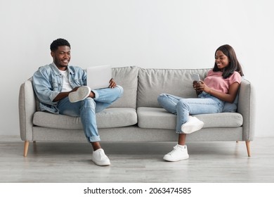 People, Technology And Lifestyle Concept. Portrait Of Happy Black Couple Sitting On Comfortable Couch In Living Room, Woman Holding Mobile Phone, Man Using Laptop Computer. Wifi Internet Connection