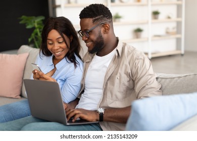 People, Technology And Lifestyle Concept. Portrait Of Happy Black Couple Sitting On Comfortable Couch In Living Room, Woman Holding Showing Mobile Phone, Man Using Pc Laptop. Wifi Internet Connection
