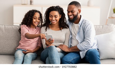 People And Technology. Portrait Of Happy African American Family Holding And Using Mobile Phone Sitting On The Couch At Home. People Spending Time Together, Sharing Social Media, Watching Funny Video