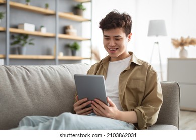 People And Technology. Portrait Of Smiling Asian Man Holding And Using Digital Tablet Sitting On Couch Indoors In Living Room. Happy Male Surfing Internet, Watching Movie Online