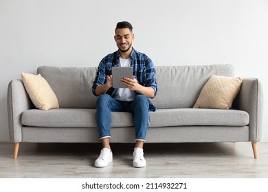 People And Technology. Portrait Of Smiling Handsome Arab Man Holding And Using Digital Tablet Sitting On Couch Indoors In Living Room. Happy Male Surfing Internet, Watching Movie Online, Choosing Gift - Powered by Shutterstock