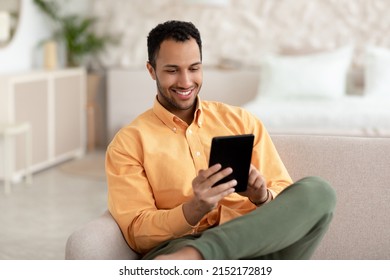 People And Technology. Portrait Of Smiling Handsome Arabic Man Holding And Using Digital Tablet Sitting On Sofa Indoors. Happy Male Surfing Internet, Reading Electronic Book, Resting In Living Room - Powered by Shutterstock