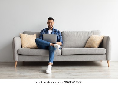 People And Technology. Portrait of young smiling Arab man holding pc on lap sitting on the sofa in living room, typing on keyboard. Cheerful guy browsing internet, surfing web, free copy space