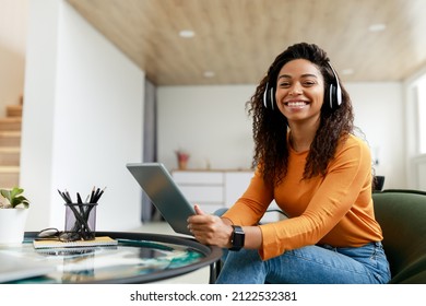 People And Technology. Portrait of young smiling black woman in wireless headset holding using digital tablet sitting on chair in living room, looking posing at camera, free copy space, banner - Powered by Shutterstock