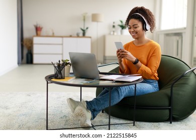 People And Technology. Smiling young black lady sitting at tea table with laptop, holding using cell phone. Cheerful female in wireless headset watching video, chatting taking break, free copy space